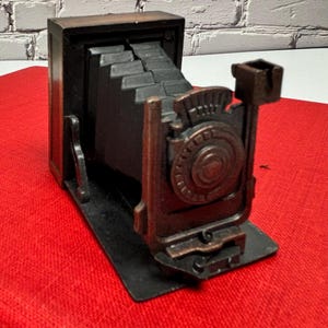 May include: A vintage-style camera figurine in shades of brown and black. The camera features a detailed front with a circular design and a bellows-style body. It sits on a black base, with a red surface in the background.
