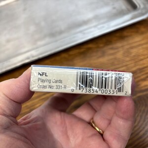 Pode incluir: Um baralho de cartas da NFL, segurado em uma m&atilde;o. A caixa de cartas &eacute; branca com detalhes em azul e vermelho. O texto "NFL Playing Cards" &eacute; vis&iacute;vel. O c&oacute;digo de barras e o n&uacute;mero do pedido tamb&eacute;m s&atilde;o vis&iacute;veis.
