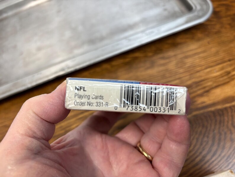 Pode incluir: Um baralho de cartas da NFL, segurado em uma m&atilde;o. A caixa de cartas &eacute; branca com detalhes em azul e vermelho. O texto "NFL Playing Cards" &eacute; vis&iacute;vel. O c&oacute;digo de barras e o n&uacute;mero do pedido tamb&eacute;m s&atilde;o vis&iacute;veis.