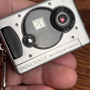 May include: A silver Innovage Mini Digital Camera with a black circular lens area, a small LCD screen displaying the number 20, and a black shutter button. The device features a metallic finish, a keychain attachment, and the text Innovage Mini Digital Camera.