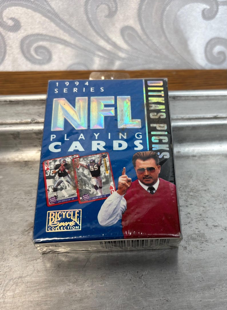 Pode incluir: Um baralho selado de cartas da NFL da s&eacute;rie de 1994. A caixa azul apresenta o logotipo da NFL, o texto "Ditka's Picks" e imagens de jogadores de futebol americano. O logotipo da Bicycle Sport Collection tamb&eacute;m &eacute; vis&iacute;vel.