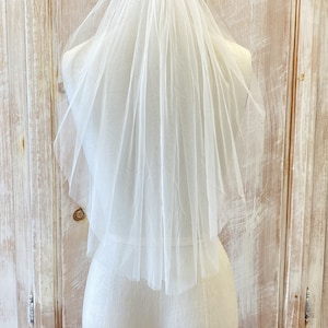 Puede incluir: Un velo de novia de tul blanco de una sola capa con un borde festoneado. El velo se muestra en un maniqu&iacute; blanco.