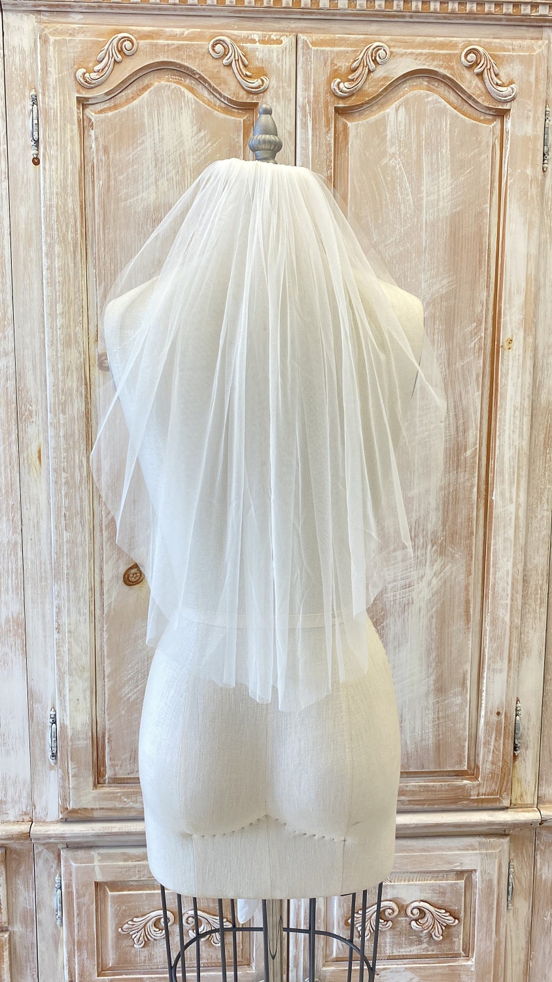 Puede incluir: Un velo de novia de tul blanco de una sola capa con un borde festoneado. El velo se muestra en un maniqu&iacute; blanco.