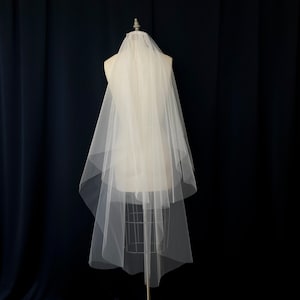 May include: A white single-tier bridal veil with a scalloped edge. The veil is made of soft tulle and is displayed on a mannequin.