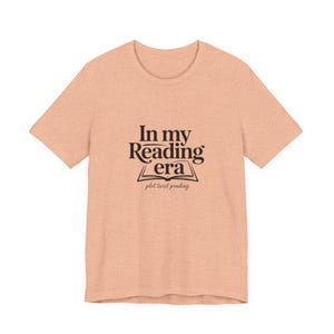 May include: A peach-coloured t-shirt featuring the text "In my Reading era" above an open book illustration, with "plot twist pending" underneath. The shirt has a crew neck and short sleeves.