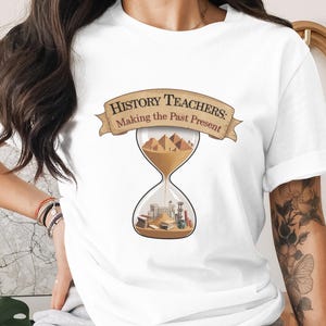 May include: White t-shirt with a graphic featuring an hourglass filled with sand and historical elements. The top of the hourglass shows pyramids, while the bottom depicts buildings and books. The text "History Teachers: Making the Past Present" is displayed above the hourglass.