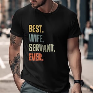 May include: Black t-shirt featuring the text "BEST. WIFE. SERVANT. EVER." in a retro, faded font. The shirt is worn by a person with tattoos on their arm and a watch on their wrist. The background is a blurred city street scene.