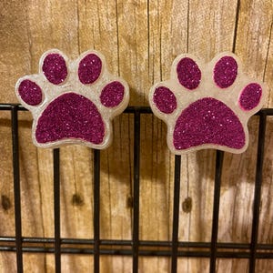 May include: Two white paw print shaped oven mitts with pink glitter. The mitts are hanging on a black wire rack.