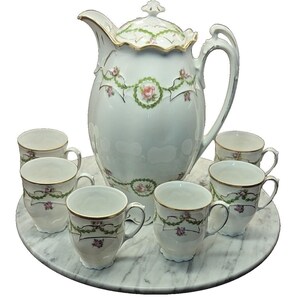 May include: A white porcelain coffee service set featuring a large lidded pitcher and five matching cups. Each piece is decorated with delicate green garlands, small pink rose motifs, and gold trim along the rims and handles.