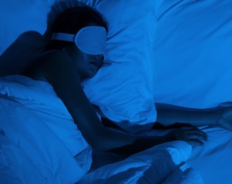 May include: A person wearing a white eye mask, lying in bed under a white blanket and pillow. The scene is illuminated with a blue light, creating a calming atmosphere. The eye mask is designed to block out light for better sleep.