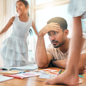 Könnte beinhalten: Ein Mann mit der Hand an der Stirn sitzt an einem Tisch mit bunten Papieren und Bleistiften. Zwei Kinder sind in Bewegung verschwommen, eines springt und der Fuß des anderen im Vordergrund, was auf eine spielerische oder chaotische Szene hindeutet.