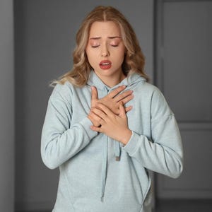 May include: A person wearing a pale blue hoodie, with hands clasped over the chest, and eyes closed. The person appears to be experiencing discomfort or pain. The background is a neutral grey.