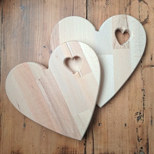 Può includere: Due taglieri di legno a forma di cuore, uno incastrato nell'altro. Il tagliere più grande ha un cuore più piccolo ritagliato al centro.