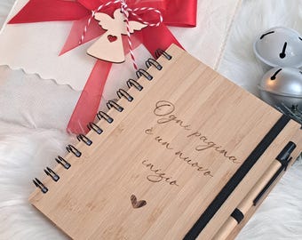 Può includere: Un taccuino in legno con il testo italiano "Ogni pagina è un nuovo inizio" e una penna, accanto a un regalo confezionato con un nastro rosso e un ornamento a forma di angelo. Il taccuino ha una rilegatura a spirale nera e una fascia elastica nera.