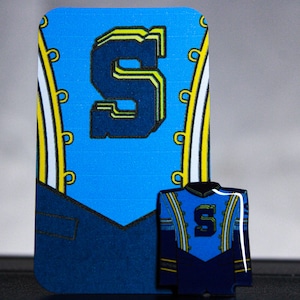 May include: A blue and navy-colored pin and a matching card featuring the letter "S". The pin is shaped like a jacket with the letter "S" on the front. The card has a large "S" and decorative elements.