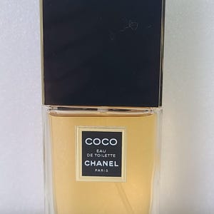 K&ouml;nnte beinhalten: Eine rechteckige Glasflasche von Coco Eau de Toilette von Chanel. Die Flasche hat einen schwarzen Deckel mit goldenem Rand und ein Etikett mit dem Markennamen und Produktdetails. Die Fl&uuml;ssigkeit im Inneren hat eine hellbernsteinfarbene Farbe.