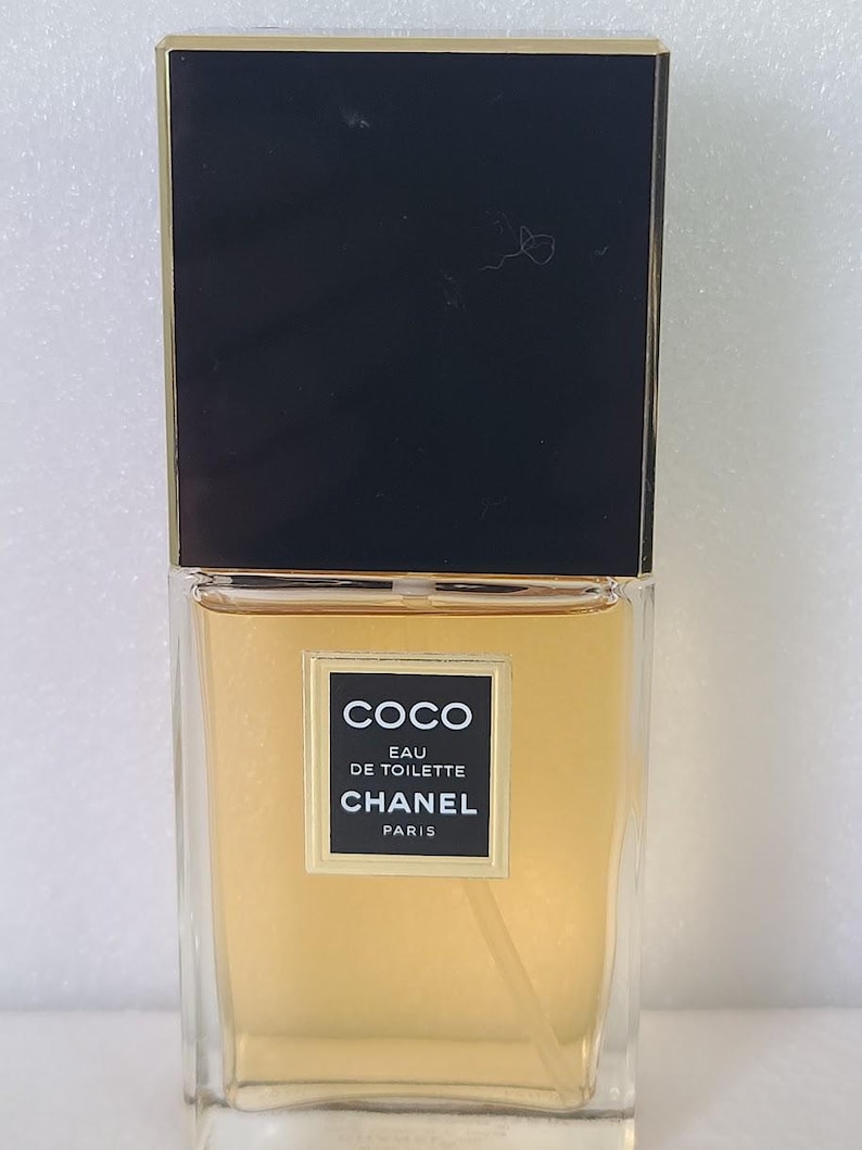 K&ouml;nnte beinhalten: Eine rechteckige Glasflasche von Coco Eau de Toilette von Chanel. Die Flasche hat einen schwarzen Deckel mit goldenem Rand und ein Etikett mit dem Markennamen und Produktdetails. Die Fl&uuml;ssigkeit im Inneren hat eine hellbernsteinfarbene Farbe.