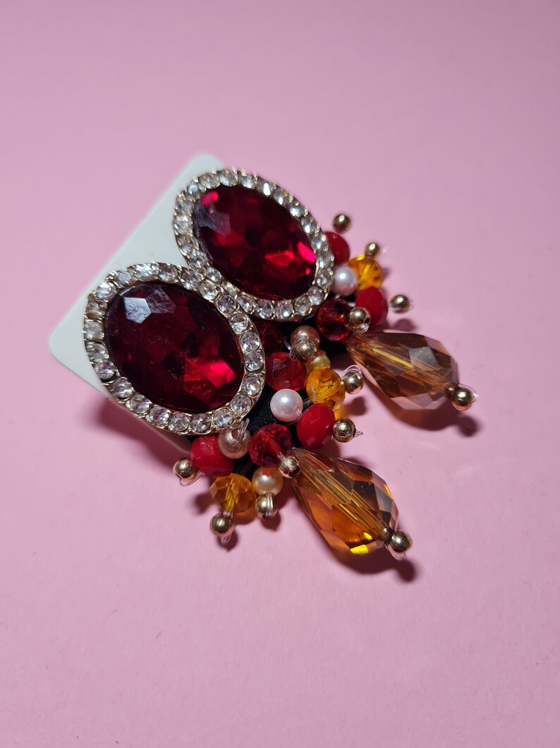 Puede incluir: Un par de pendientes rojos y dorados con cristales colgantes. Los pendientes presentan una gran piedra roja rodeada de peque&ntilde;os diamantes de imitaci&oacute;n transparentes. Los cristales colgantes son &aacute;mbar y rojos.
