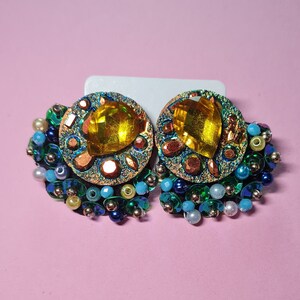 Puede incluir: Un par de pendientes llamativos con un gran cristal amarillo rodeado de lentejuelas y cuentas iridiscentes en verde, azul y dorado.