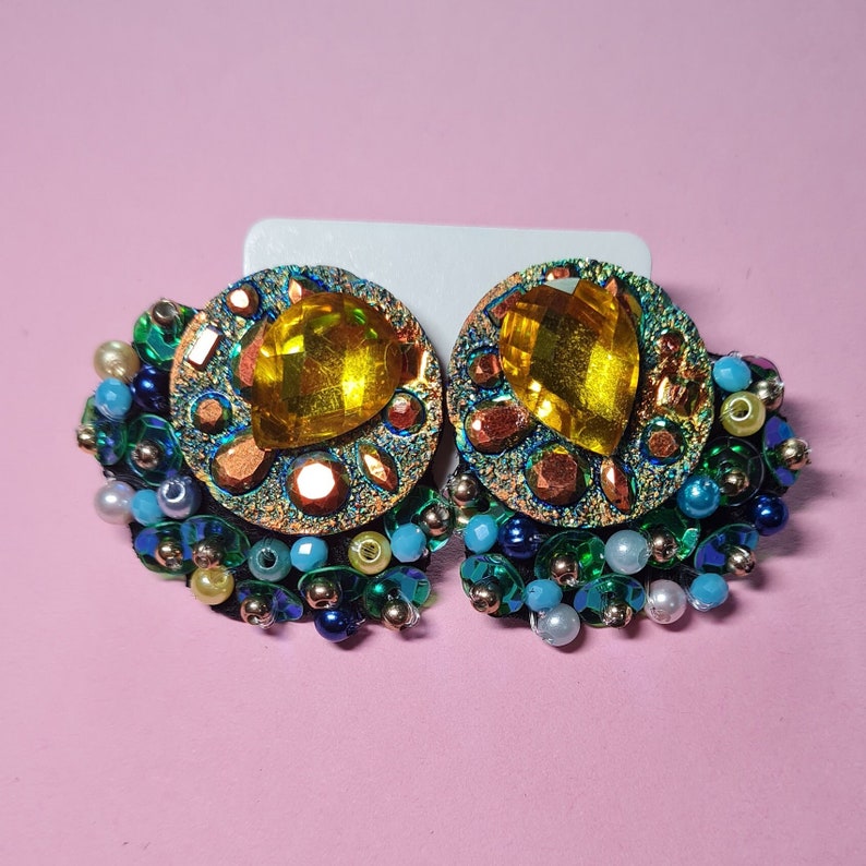 Puede incluir: Un par de pendientes llamativos con un gran cristal amarillo rodeado de lentejuelas y cuentas iridiscentes en verde, azul y dorado.