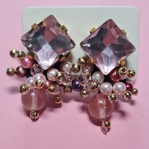 Puede incluir: Un par de pendientes de cristal rosa con detalles dorados y un racimo de cuentas rosas, blancas y doradas.