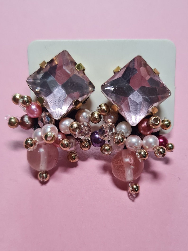 Puede incluir: Un par de pendientes de cristal rosa con detalles dorados y un racimo de cuentas rosas, blancas y doradas.