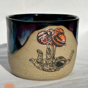 Puede incluir: Taza de cerámica con exterior beige moteado y borde azul oscuro y morado. La taza presenta una ilustración en blanco y negro de un astronauta flotando con globos con forma de planeta.