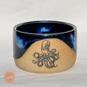 May include: A handmade ceramic cup with a dark blue glazed rim and a tan base. A black octopus illustration is centered on the cup's exterior. The cup is cylindrical and appears to be a small bowl or mug.