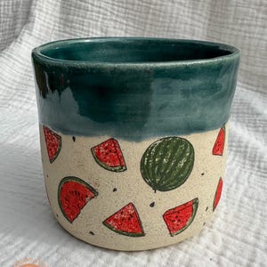 Puede incluir: Una taza de cerámica con borde turquesa y cuerpo beige decorada con rodajas de sandía y una sandía entera. La taza es cilíndrica y parece hecha a mano, con una estética rústica. El diseño de sandía presenta colores rojos y verdes.
