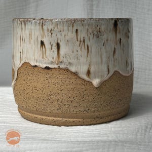 May include: A handcrafted ceramic planter with a speckled, tan base and a creamy white glaze that drips down the sides. The planter has a cylindrical shape and a rustic, earthy aesthetic. The planter is suitable for indoor or outdoor use.