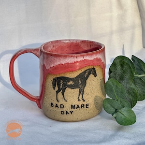 Peut inclure: Une tasse en céramique avec un bord et une anse rouges et blancs. La tasse présente une illustration de cheval noir et le texte "BAD MARE DAY" en noir. La tasse a une base beige.
