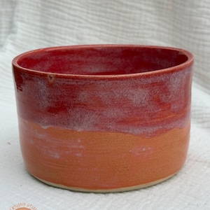 May include: A handmade ceramic bowl with a gradient of colors. The top is a deep red, fading to a lighter red and then to a warm orange at the base. The bowl has a cylindrical shape and a smooth finish. The logo "Sterlings Studio" is visible.