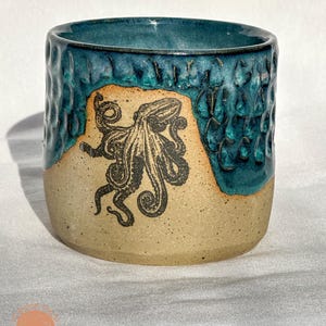 May include: A handmade ceramic mug with a teal blue glazed rim and exterior. The lower portion is a tan color with a black octopus illustration. The mug has a textured surface.