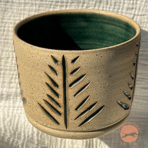 May include: A speckled beige ceramic planter with a dark green interior. The exterior features a carved leaf design and a rounded base. The planter is suitable for indoor or outdoor use.