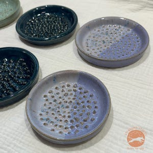 May include: A collection of handmade ceramic soap dishes in various shades of blue and gray. Each dish is round with a textured surface of small, raised bumps for soap drainage. The dishes are glazed with a glossy finish.