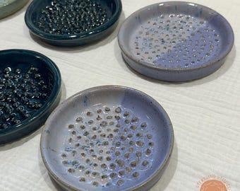 Pode incluir: Uma coleção de saboneteiras de cerâmica feitas à mão em vários tons de azul e cinza. Cada prato é redondo com uma superfície texturizada de pequenas saliências elevadas para a drenagem do sabão. Os pratos são esmaltados.
