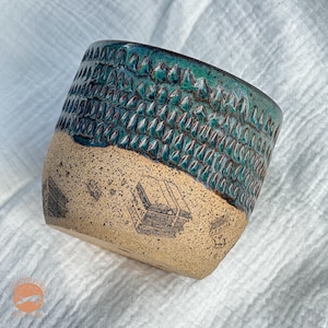 May include: Handmade ceramic mug with a textured teal glaze on the upper half and a tan, speckled base. The base features black illustrations of books. The mug is designed for beverages.