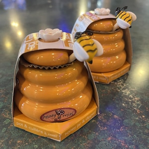 May include: Two ceramic honey pots shaped like beehives, with a yellow and black bee figurine on top. The pots are orange with a white flower on the lid. The packaging reads "honey miel" and "honey jar & dipper".
