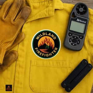 Puede incluir: Una camisa de trabajo amarilla con un parche de "Wildland Firefighter", guantes de cuero, un medidor meteorológico Kestrel y una multiherramienta negra. El parche presenta una escena de montaña con llamas y el texto "Wildland Firefighter".