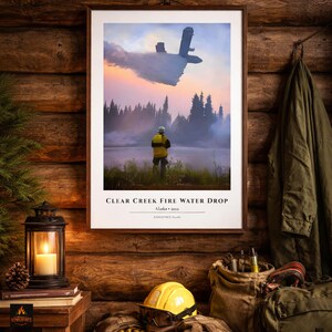 以下が含まれることがあります： 「CLEAR CREEK FIRE WATER DROP」と題された額入りのプリントは、消防士がいる湖の上を飛ぶ散水飛行機を描いています。画像は夕焼けと森を背景に設定されています。ランタン、ヘルメット、ギアが前景にあります。