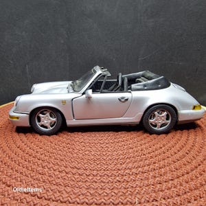 May include: A silver Porsche 911 convertible model car with a black top. The car is on a brown textured surface.