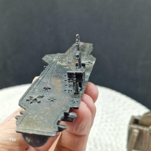 May include: A detailed, miniature metal aircraft carrier model in shades of gray and brown. The model features intricate details of the ship's structure, including the flight deck, control tower, and other components. The model is held in a hand.
