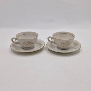 May include: Two white teacups with gold trim and floral designs on the side, each with a matching saucer.