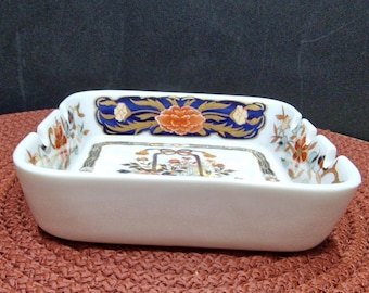 May include: Square white ceramic ashtray with a blue, gold, and red floral design. The ashtray has a raised rim and four indentations for cigarettes.