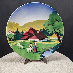 Op de afbeelding: Een decoratieve keramische schotel met een landelijke landschapsscène. De schotel toont een huis met een rood dak, groene velden, een beek en figuren van mensen en geiten. De schotel wordt ondersteund door een houten standaard en heeft een blauwe lucht op de achtergrond.