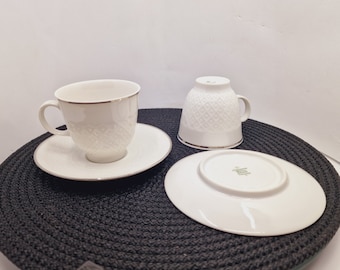 May include: Two white porcelain teacups and saucers with a delicate embossed pattern and a silver trim. The cups and saucers are on a black round woven placemat.