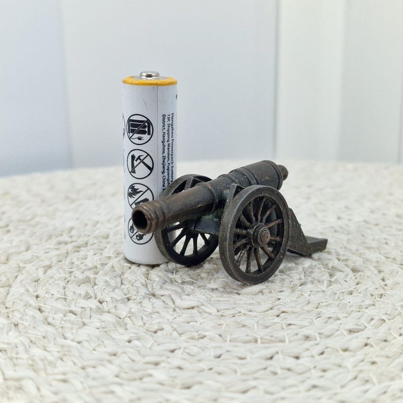 May include: A miniature antique-style cannon made of dark metal, with spoked wheels and a detailed barrel. Next to it is a white AA battery with black symbols and text. The cannon is on a textured, off-white surface.