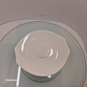 Może przedstawiać: Biały spodek ceramiczny z lekko zakrzywionym brzegiem i małym wgłębieniem w środku. Spodek znajduje się na przezroczystej powierzchni szklanej.