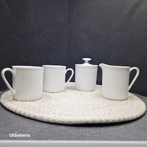 Puede incluir: Un juego de cuatro tazas de cerámica blanca y una jarra de crema con un diseño acanalado. Las tazas y la jarra de crema están dispuestas sobre un mantel tejido beige.