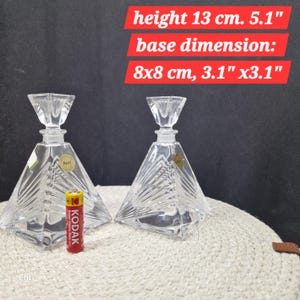 May include: Two clear crystal perfume bottles with geometric pyramid shapes. Each bottle has a height of 13 cm and a base dimension of 8x8 cm. The bottles have a decorative stopper and are labeled "Bari".